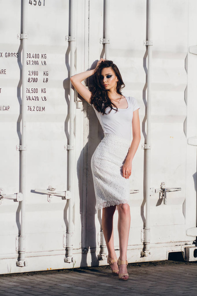 Young beautiful brunette girl with long hair in elegant white dress. Woman walking through the streets of the city in the evening at sunset. - Foto, Imagem