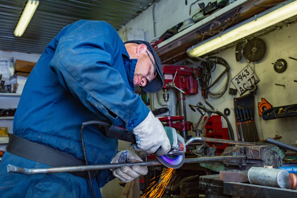 Un giovane saldatore in una t-shirt blu, occhiali e guanti da costruzione elabora metallo una smerigliatrice angolare in garage, sullo sfondo un sacco di strumenti
 - Foto, immagini