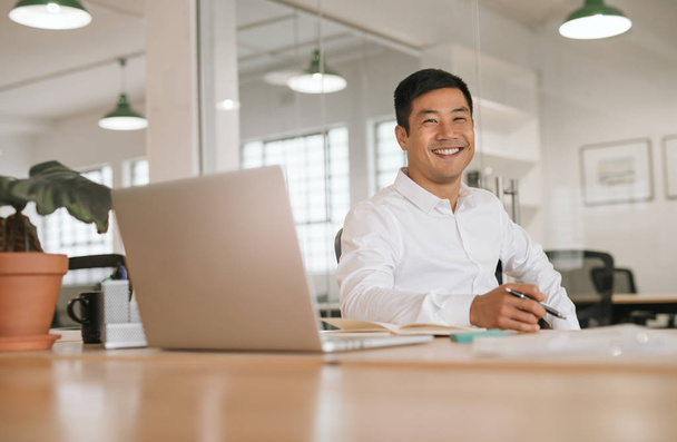 Portrait of a smiling young Asian businessman sitting at his desk in an office going over paperwork and using a laptop - Foto, immagini