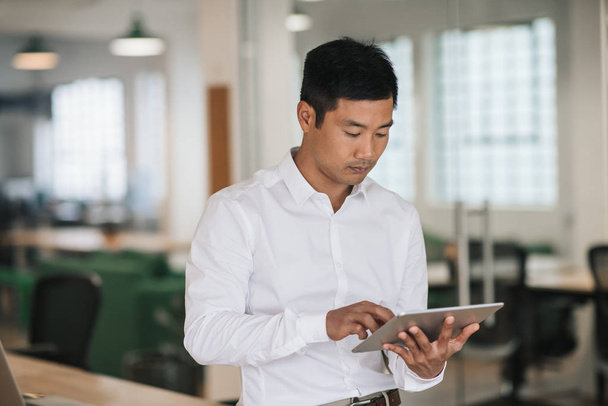 Focado jovem empresário asiático trabalhando on-line com um tablet digital, enquanto está sozinho em um grande escritório moderno
 - Foto, Imagem