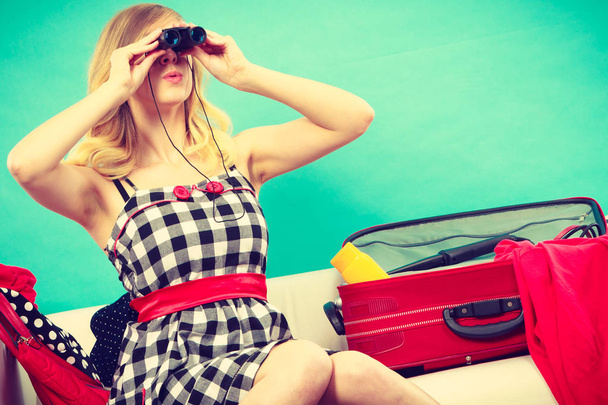 Packing for journay, traveling concept. Woman looking through binoculars, sitting on couch with suitcase and full of things ready to be taken on summer holiday. - Photo, Image