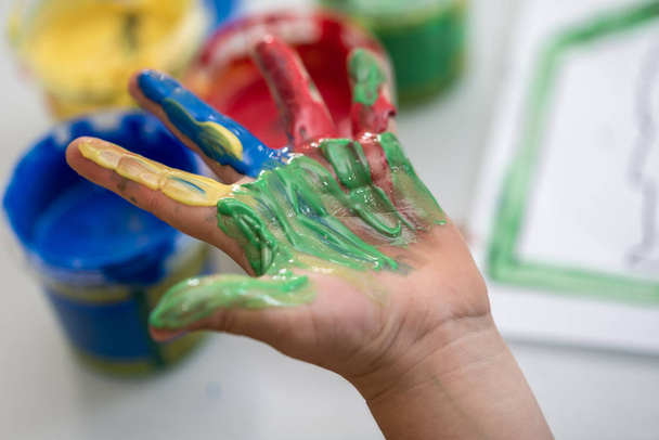 Happy artistic child showing off a colourful hand painted with red, yellow, green and blue hand paints during a kindergarten art class in a close up view. - 写真・画像