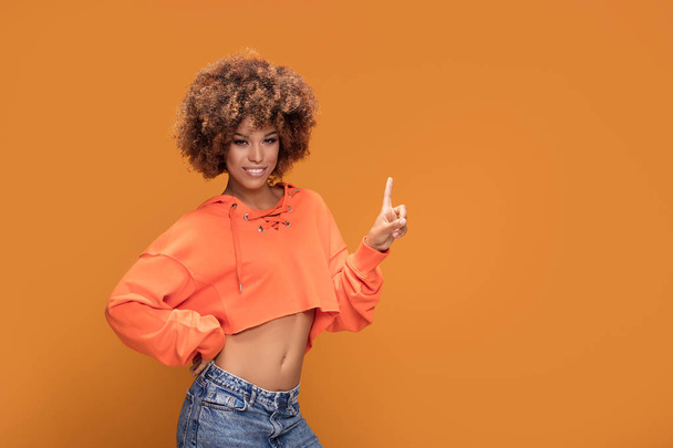 Photo of an african american woman with glamour makeup and afro hairstyle dressed in fashionable short orange blouse. Girl looking at camera showing empty yellow background. - Foto, imagen