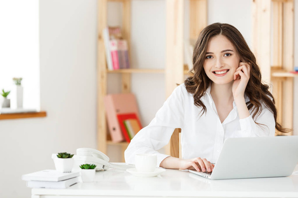 Χαμογελώντας νεαρή γυναίκα που κάθεται μπροστά από το φορητό υπολογιστή στο γραφείο. - Φωτογραφία, εικόνα