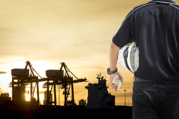 Factory engineer concept, Back view of foreman holding helmet and silhouette industrial shipping port and sunrise, transportation import export - Photo, Image