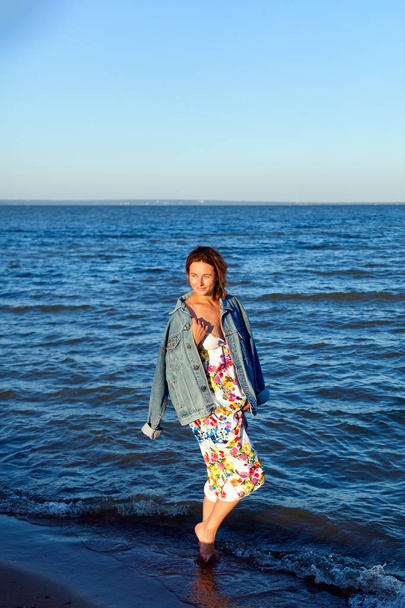 Uma mulher alegre de cabelos escuros no fio colorido e jaqueta de ganga sorri, caminha ao longo da praia e desfruta do sol brilhante em um dia de verão
 - Foto, Imagem