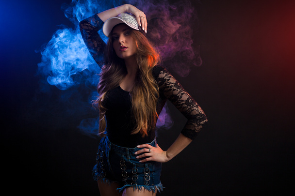 Young dark-haired woman in black top, jeans and cap posing against a background of  blue smoke from a vape on a black isolated background - Foto, imagen