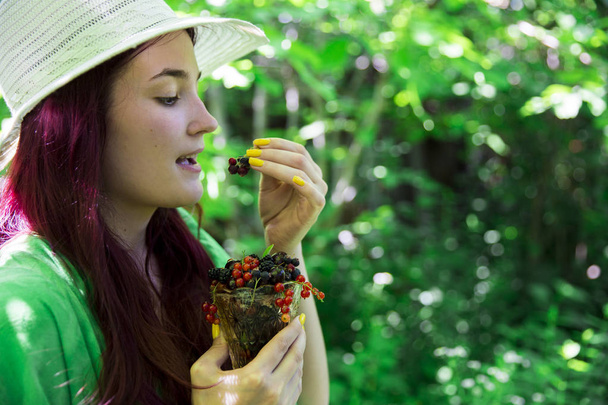 Im Profil isst ein junges Mädchen mit Strohhut eine Kirsche und hält in der Hand ein Glas mit reifen aromatischen Beeren von schwarzen und roten Johannisbeeren, Kirschen, Kirschen und Brombeeren auf einem verschwommenen Hintergrund des Gartens - Foto, Bild