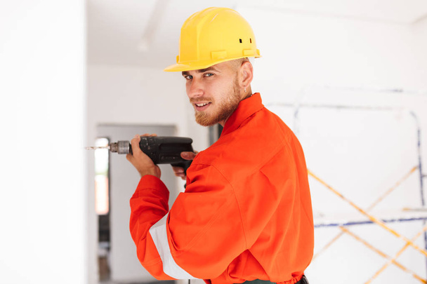 Jeune contremaître en vêtements de travail orange et casque jaune regardant à la caméra à l'aide d'une perceuse au travail
 - Photo, image