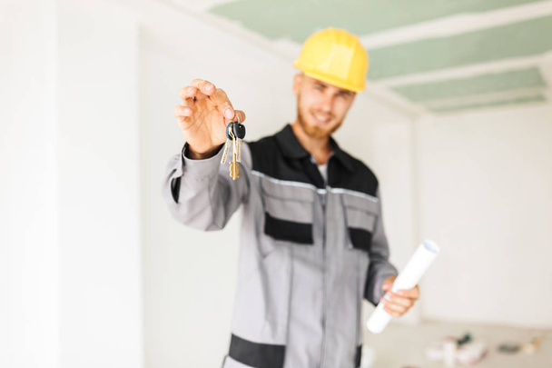 Nahaufnahme Foto des jungen Ingenieurs in Arbeitskleidung und gelbem Bollenhut mit den Schlüsseln der neuen Wohnung in der Hand  - Foto, Bild
