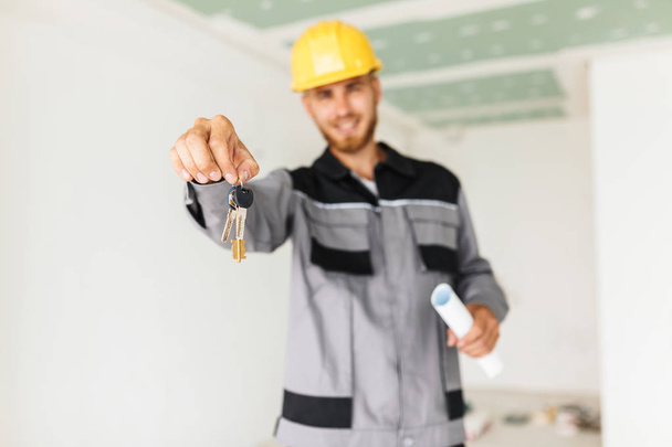 Ingeniero de cerca en ropa de trabajo y hardhat amarillo felizmente sosteniendo las llaves de nuevo apartamento en la mano
  - Foto, Imagen
