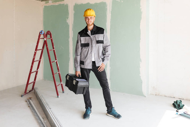 Улыбающийся инженер в рабочей одежде и в жёлтой каске с инструментами в руке счастливо смотрит в камеру с красной лестницей на заднем плане
 - Фото, изображение