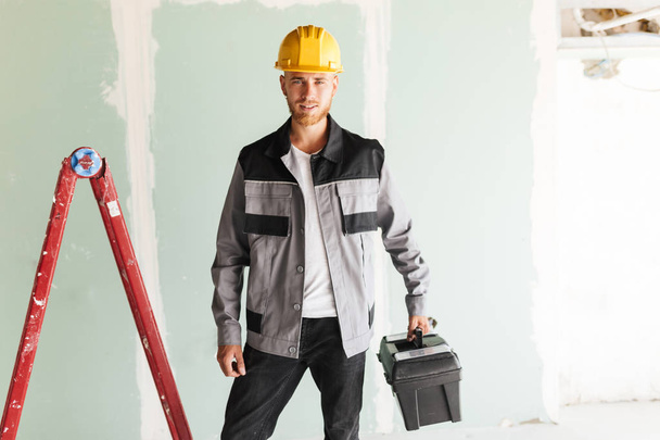 Молодой улыбающийся инженер в рабочей одежде и в жёлтой каске с инструментами в руке мечтательно смотрит в камеру с красной лестницей на заднем плане
 - Фото, изображение