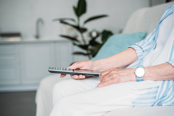 обрезанный снимок пожилой женщины с дистанционным управлением, смотрящей телевизор на диване дома
 - Фото, изображение