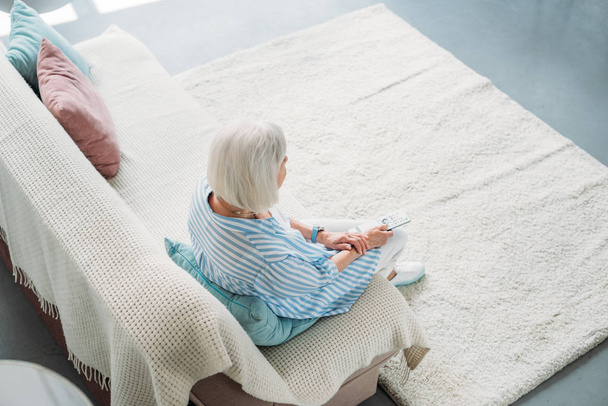 pohled z vysokého úhlu starší ženy s dálkovým ovládáním, sledování televize na gauči doma - Fotografie, Obrázek