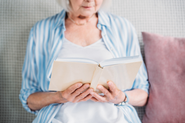 cropped shot of senior woman reading book while resting on sofa at home - 写真・画像