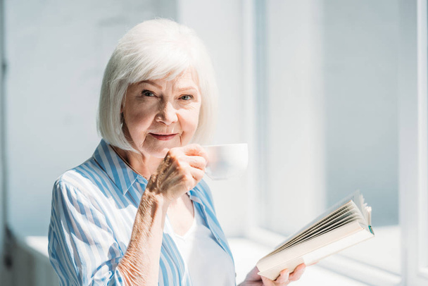 side view of grey hair woman with book and cup of coffee standing at windowsill at home - Foto, Bild