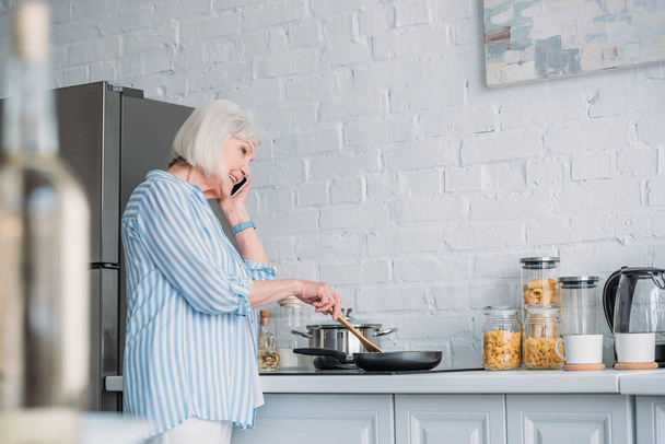 vista laterale della donna anziana sorridente che parla sullo smartphone mentre cucina la cena in cucina
 - Foto, immagini