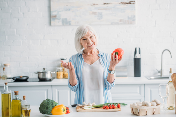 Portret van glimlachen grijs haar dame met paprika in de hand permanent aan balie in keuken - Foto, afbeelding