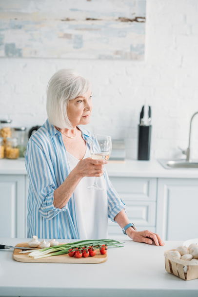 pensive senior lady with glass of wine standing at counter with fresh vegetables on cutting board in kitchen - 写真・画像