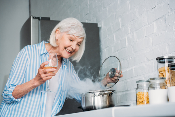 Вид сбоку улыбающейся пожилой дамы с бокалом вина, проверяющей соус на плите на кухне
 - Фото, изображение
