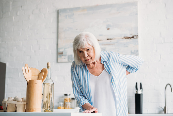 Portret van senior dame met rugpijn leunend op de teller in de keuken - Foto, afbeelding