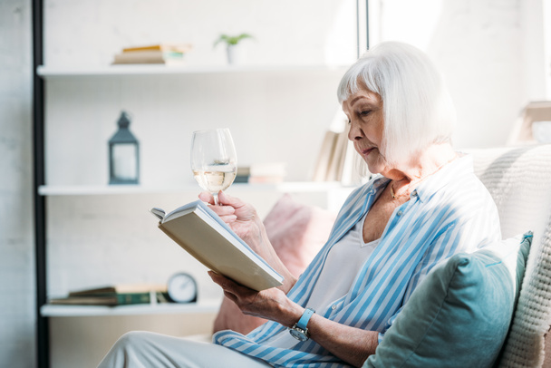 vue latérale de la femme âgée concentrée avec un verre de vin livre de lecture sur le canapé à la maison
 - Photo, image
