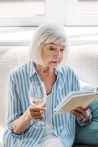 portrait de femme âgée concentrée avec verre de vin livre de lecture sur le canapé à la maison
 - Photo, image
