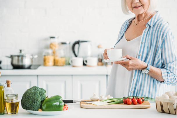 tiro recortado de mujer mayor con taza de café de pie en el mostrador con verduras frescas para la cena en la cocina
 - Foto, Imagen