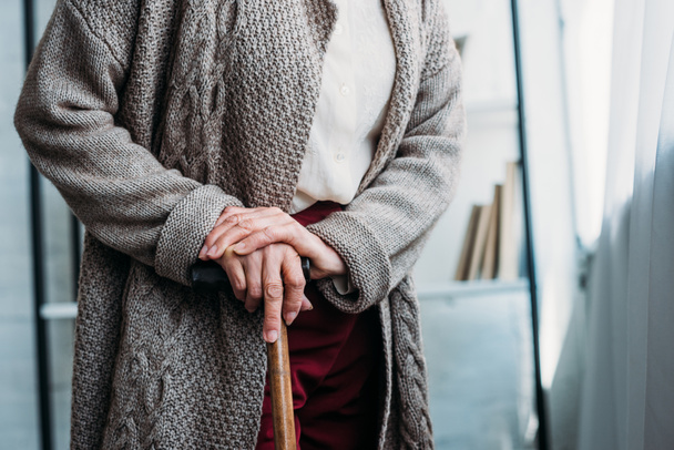 cropped shot of senior lady with wooden walking stick standing at home - Photo, Image