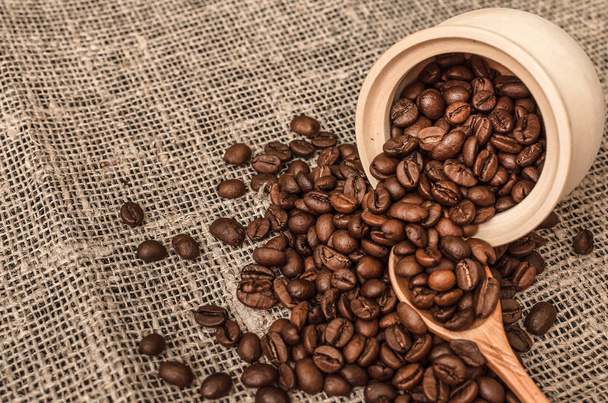 Coffee beans scattered from a wooden pot and wood spoon on burlap cloth background. Close up photo. - Photo, image