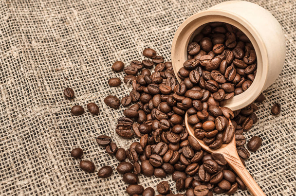 Coffee beans scattered from a wooden pot and wood spoon on burlap cloth background. Close up photo. - Valokuva, kuva