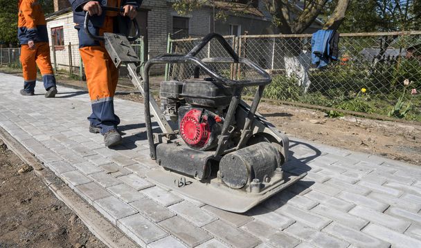 Hombre en uniforme naranja usando la máquina de piedra de pavimentación vibracional para el acabado en un sitio de construcción de carreteras en la acera
. - Foto, Imagen