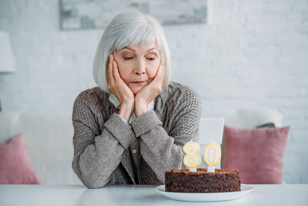 pensativo señora mayor sentado a la mesa con pastel de cumpleaños con velas solo en casa
 - Foto, imagen