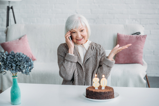 portrait of happy senior woman talking on smartphone while sitting at table with birthday cake at home - Foto, Bild