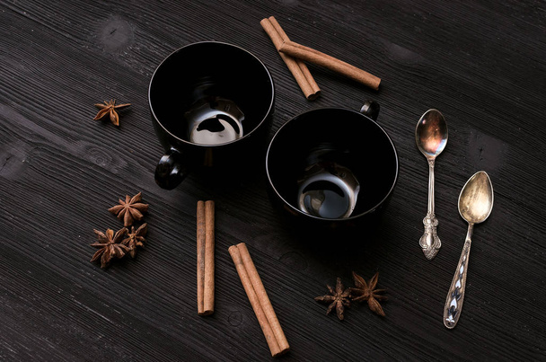 Two empty black tea cup and star anise with cinnamon and set of silver spoon around on black wooden table background. - 写真・画像