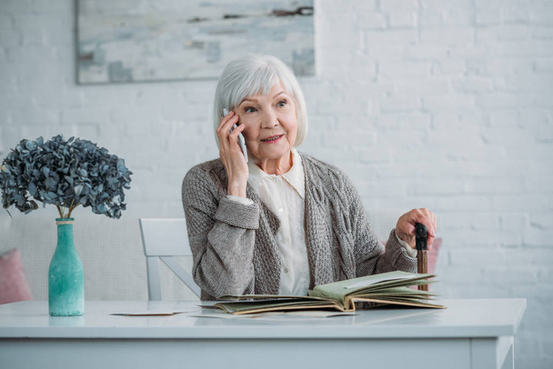 Porträt einer Seniorin mit Gehstock, die am Smartphone spricht, während sie zu Hause mit Fotoalbum am Tisch sitzt - Foto, Bild