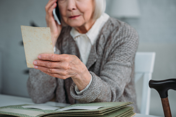 Teilbild einer Seniorin mit Foto in der Hand, die zu Hause mit dem Smartphone spricht - Foto, Bild