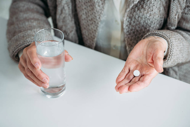 partial view of senior woman with pill and glass of water in hands sitting at table alone at home - Foto, immagini
