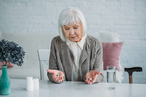 Porträt einer grauhaarigen Frau mit Tabletten in den Händen, die allein zu Hause am Tisch sitzt - Foto, Bild