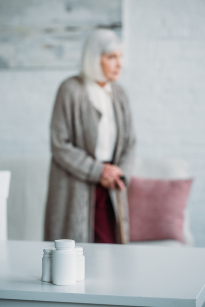 selective focus of senior woman with walking stick standing in room with medicines on tabletop at home - Foto, immagini