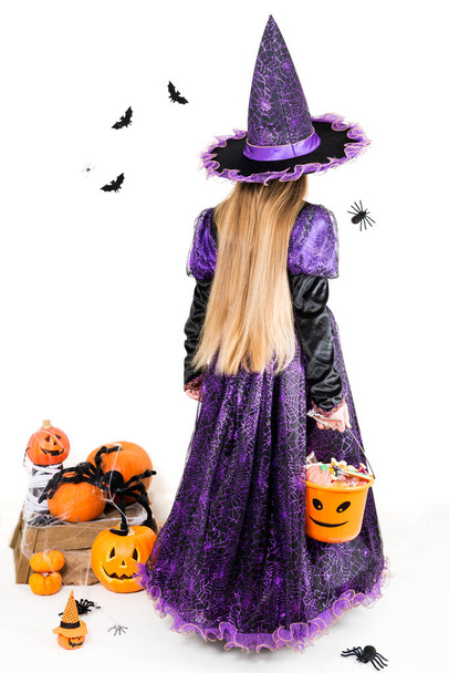 Mädchen in einem Hexenkleid mit Halloween-Accessoires und einem vollen Eimer voller Süßigkeiten auf isoliertem Hintergrund - Foto, Bild