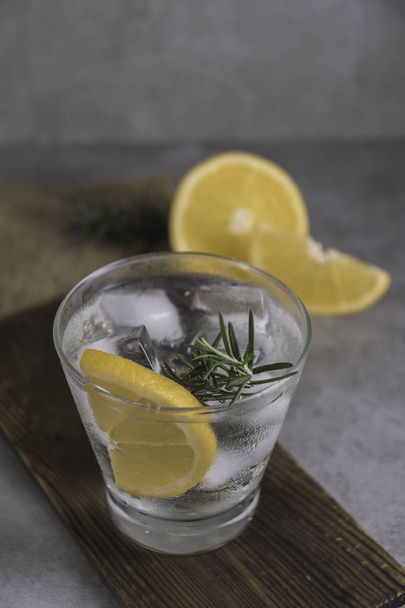 Food composition of gin tonic cocktail with lemon, rosemary and ice on table. - Photo, image