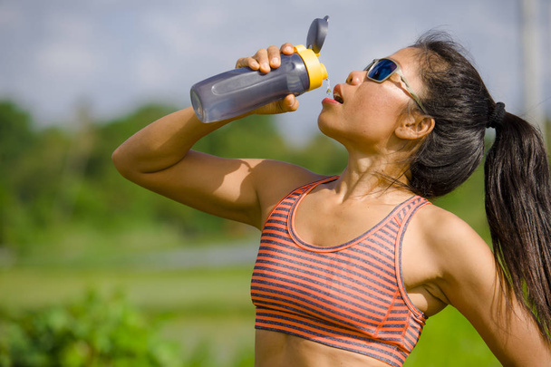 attractive and fit Asian runner woman holding isotonic bottle drinking water after training and running series workout at outdoors track in green field background in hydration and recovery  - Foto, Imagem
