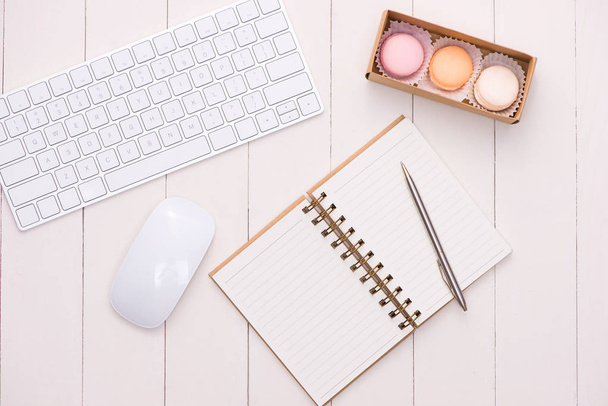 Λευκό γραφείο με πολύχρωμα μακαρόν, πληκτρολόγιο και ανοίξτε το Σημειωματάριο. - Φωτογραφία, εικόνα