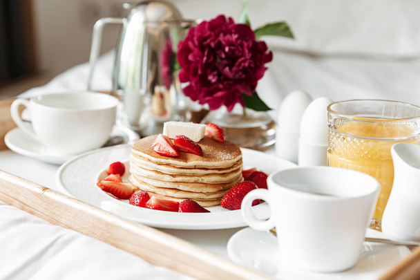 Breakfast in bed with white linen. Pancakes, boiled eggs, coffee, orange juice on tray at hotel apartment or home interior - Photo, Image