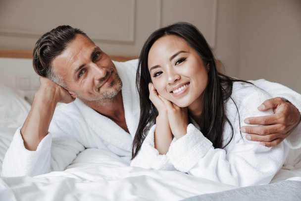 Happy young couple dressed in bathrobes lying on bed and hugging - Фото, изображение