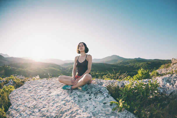 Tyttö istuu vuoren huipulla auringonlaskun aikaan. Nainen harrastaa joogaa. Meditaatio on kaukana sivilisaatiosta. Kauniin luonnon mietiskelyä. Näkymä vuorille
. - Valokuva, kuva