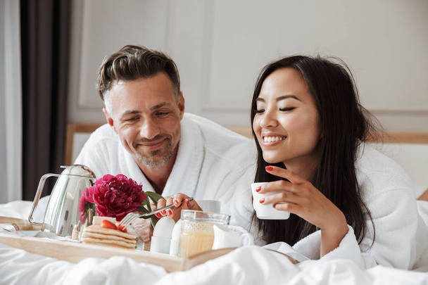 Photo of beautiful asian woman in white housecoat eating strawberry and pancakes while having breakfast in bed during vacation with handsome man at hotel apartment - Zdjęcie, obraz