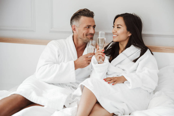Beautiful couple 30s caucasian man and asian woman wearing white housecoat smiling and drinking champagne from glasses while lying in bed at hotel apartment - Fotoğraf, Görsel